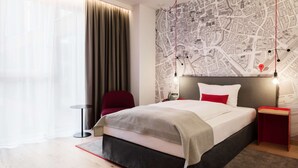A modern hotel room with a large bed, a red chair, a small table, and a map wall. - IntercityHotel Braunschweig (Braunschweig)