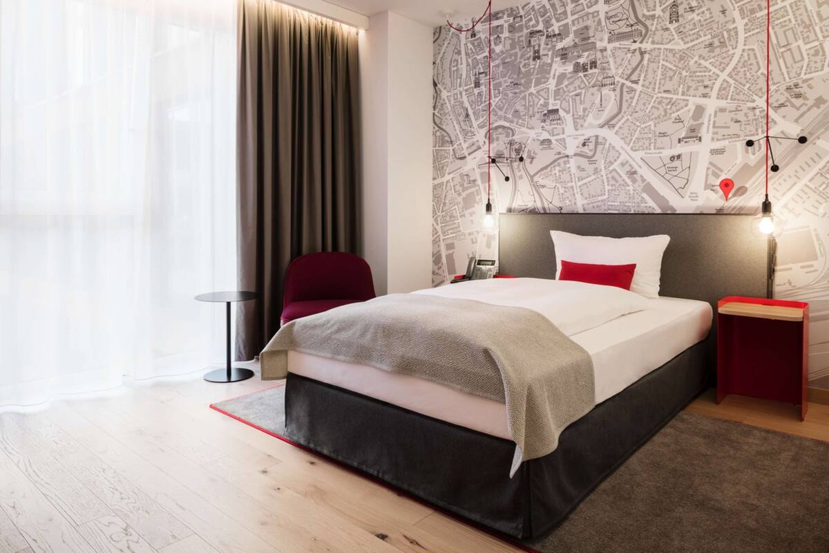 Quarto de hotel moderno com uma cama grande, uma cadeira vermelha, uma mesa pequena e uma parede com mapa.