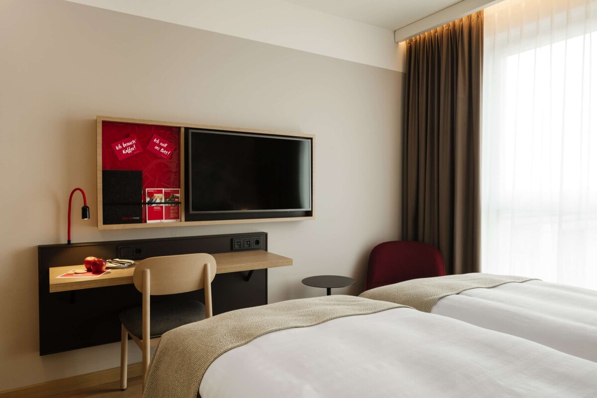 A hotel room with a bed, a desk with a chair, a television, and a window with curtains.