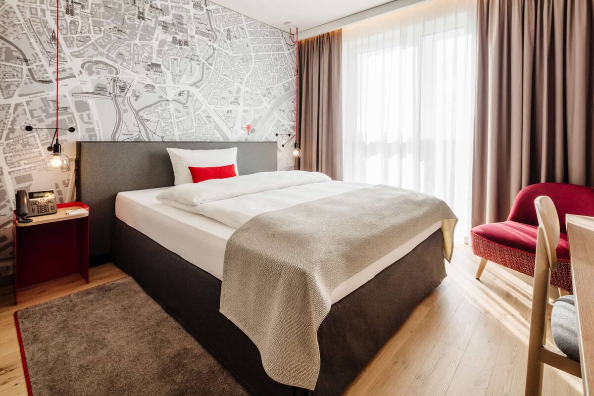 Quarto de hotel com uma cama grande, uma cadeira vermelha, um mural com mapa e um telefone vermelho no criado-mudo.