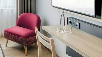 A modern hotel room with a wooden desk, a red upholstered chair, a glass table, and a large window.