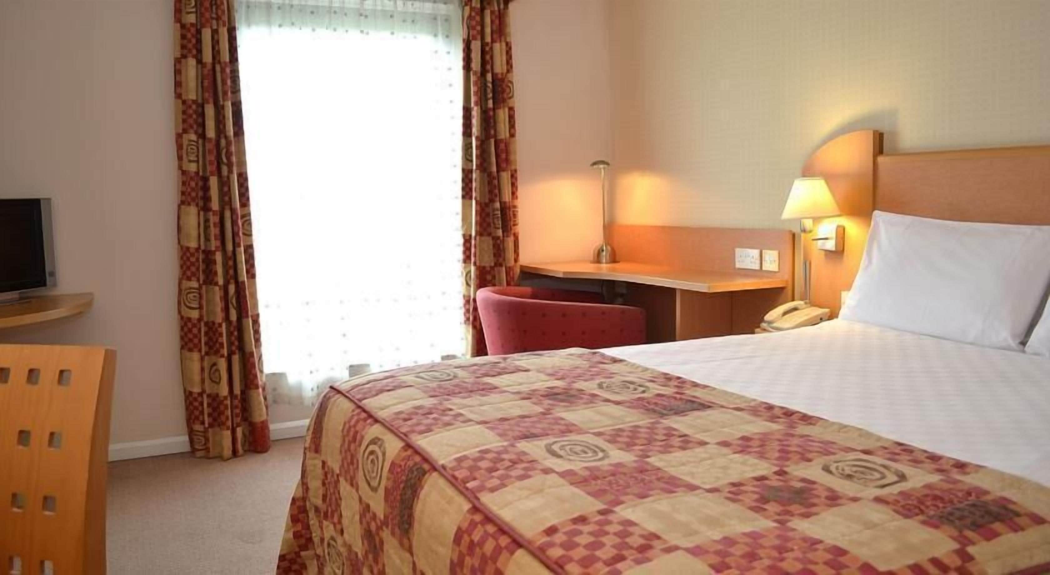 A hotel room with a bed, a desk, a chair, a TV, and a telephone.