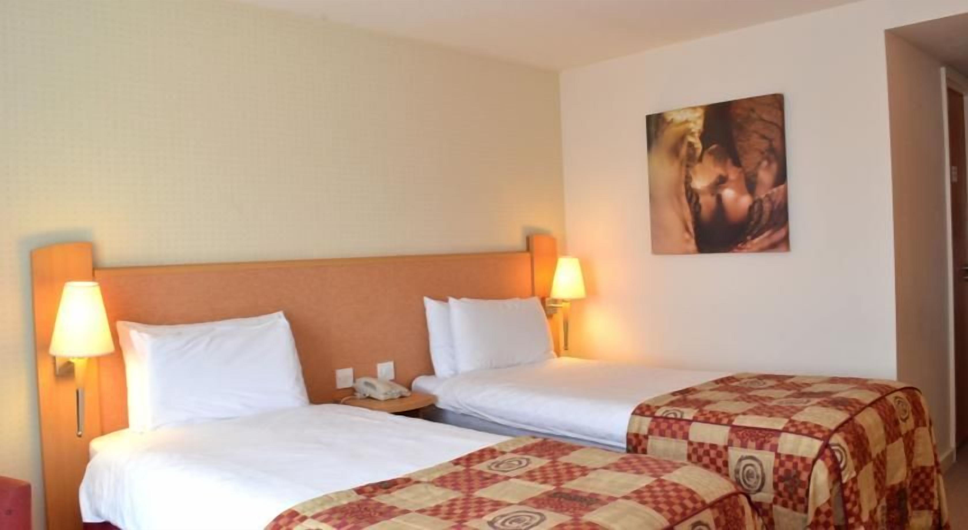 A hotel room with two beds, a nightstand, a lamp, and a painting on the wall.