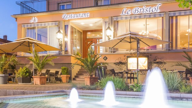 A two-story building with a restaurant named "Le Bristol" on the ground floor. There is a fountain in front of the building, and outdoor seating with umbrellas. - Hotel Restaurant Le Bristol (Niederbronn-les-Bains)