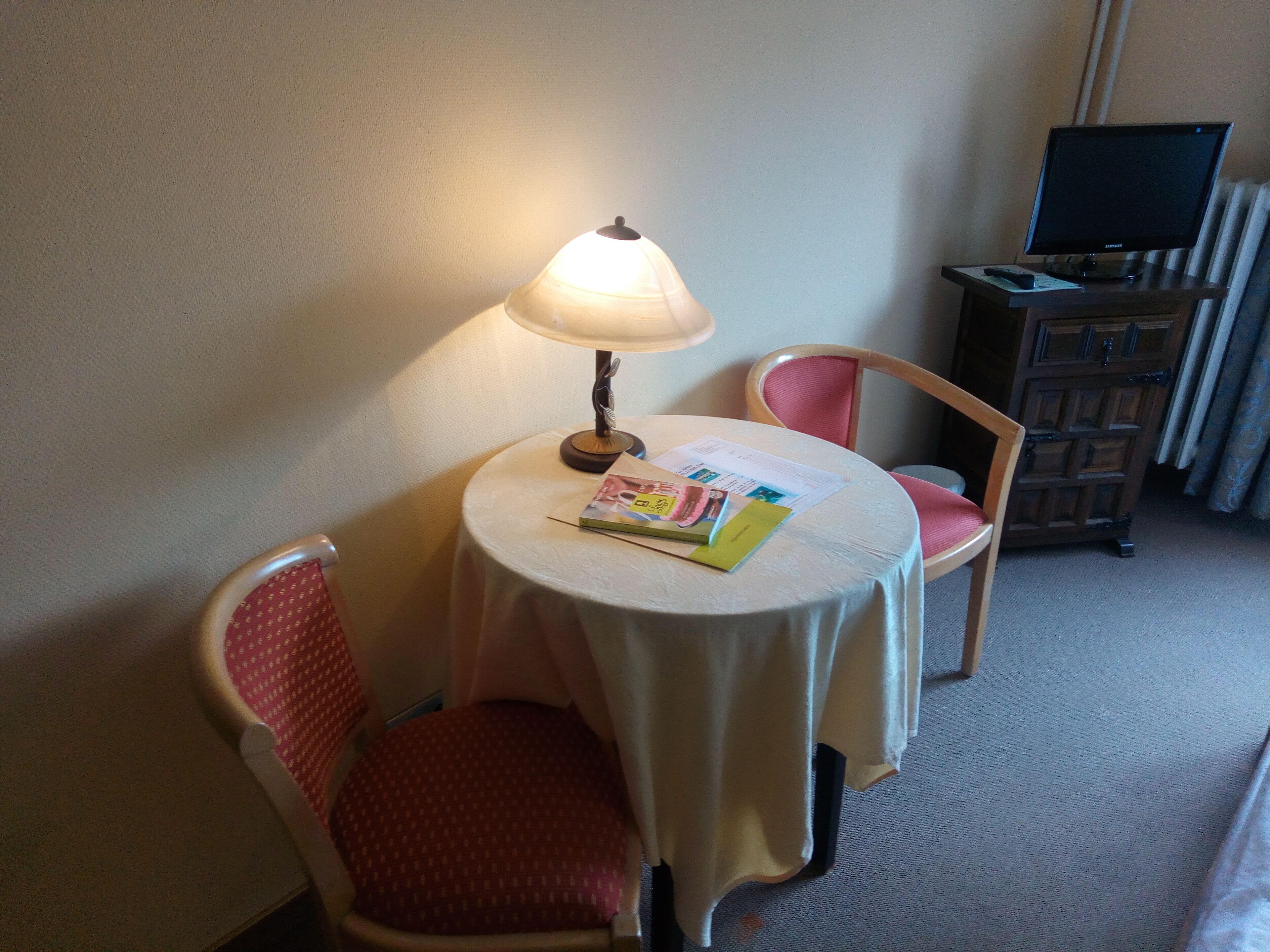 A small, cozy room with a round table, a lamp, a chair, and a television on a stand.