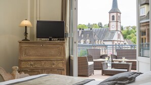 A hotel room with a balcony, a television on a wooden dresser, wicker furniture, and a view of a church with a clock tower. - Hotel Restaurant Le Bristol (Niederbronn-les-Bains)
