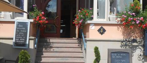 A building entrance with a staircase leading to a door, flower boxes with red flowers, and two signs with text.