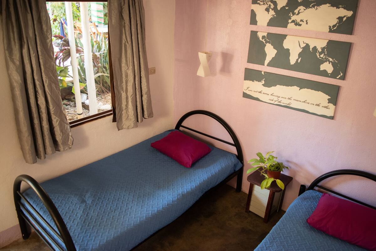 A small, single-bed room with a window, a wall-mounted world map, and a small table with a plant.