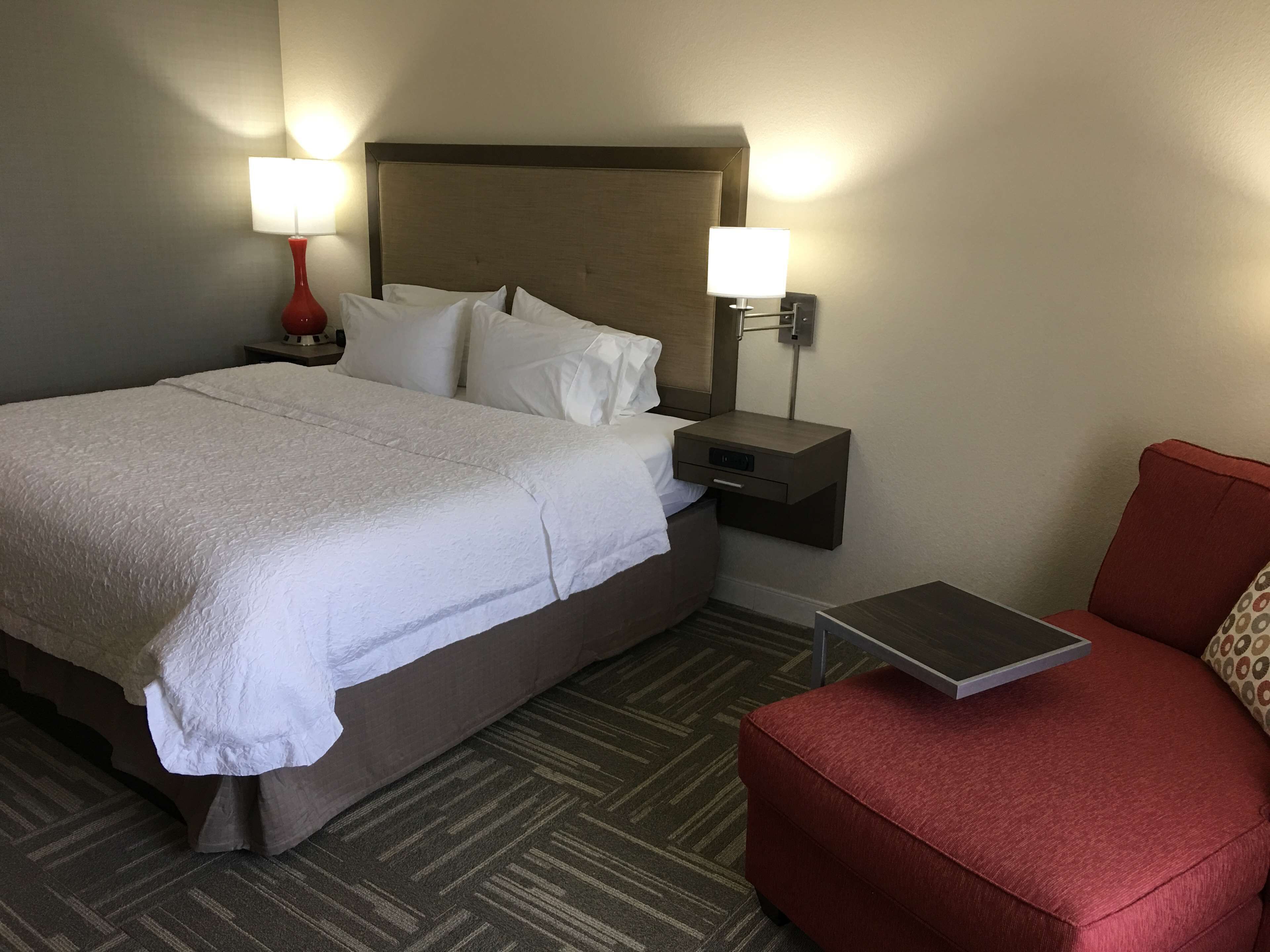 A hotel room with a large bed, two bedside lamps, a nightstand, a red chair, and a small table.