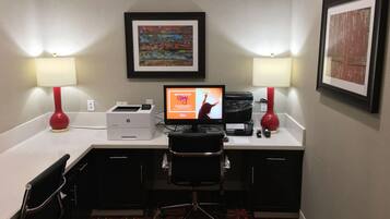 A hotel room with a desk, two lamps, a printer, a computer, a chair, and a carpet.