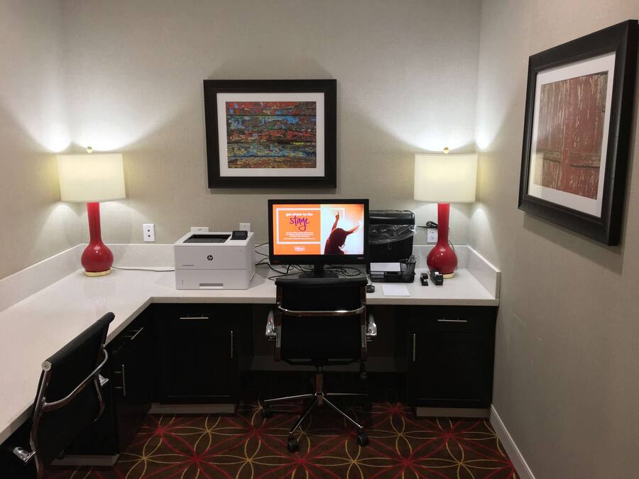 A hotel room with a desk, two lamps, a printer, a computer, a chair, and a carpet.