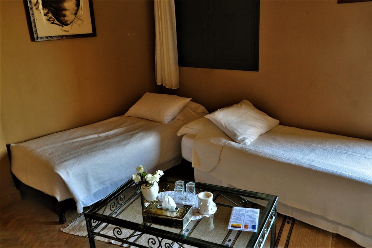 A hotel room with two single beds, a small table with a tray, a vase with flowers, and a framed picture on the wall.