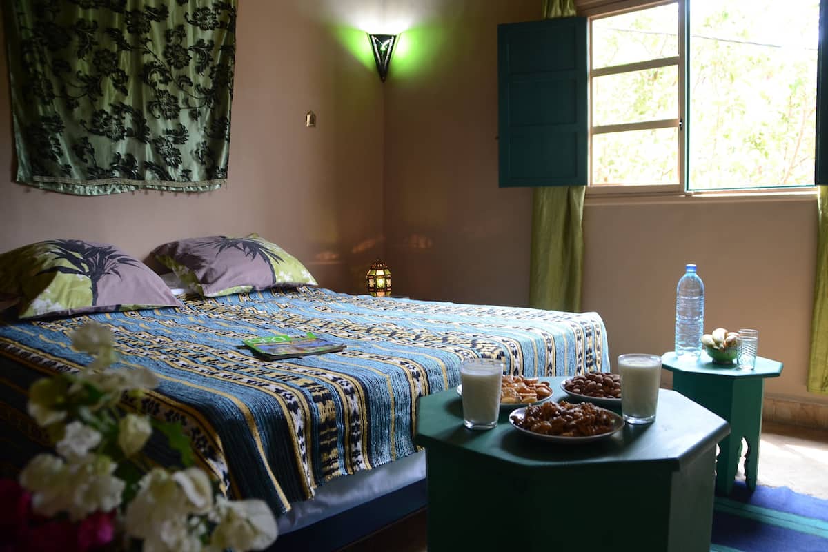 A bedroom with a bed, bedside table with food, a window, and a wall hanging.
