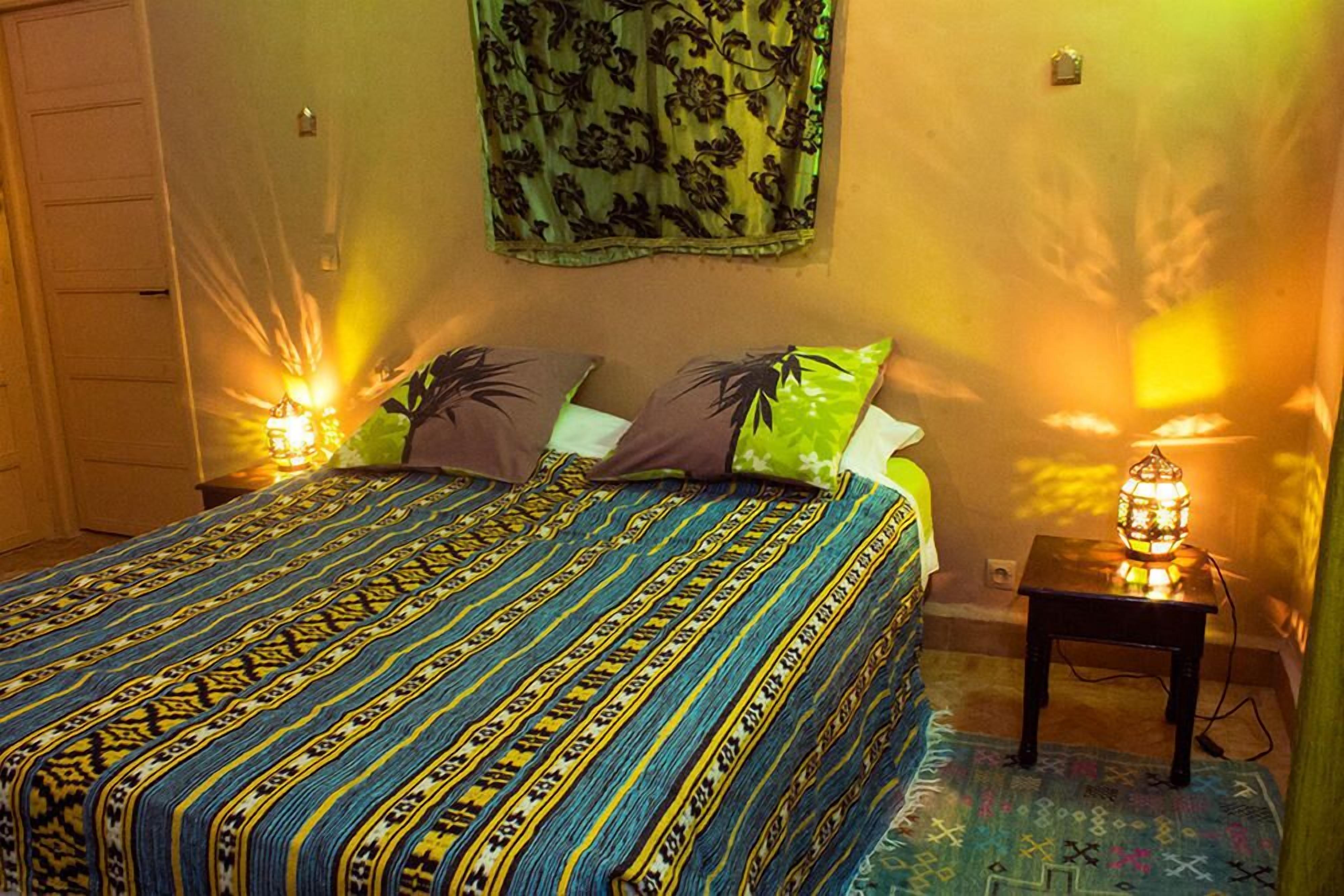 Un dormitorio con una colcha estampada, dos almohadas, un tapiz en la pared y una lámpara de noche.