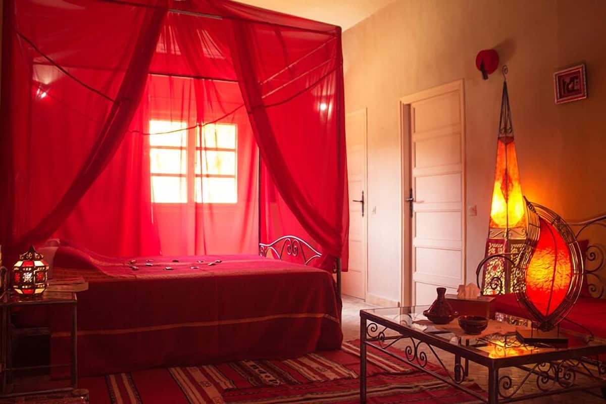 A bedroom with a bed under a red canopy, a bedside table with a lamp, and a door in the background.