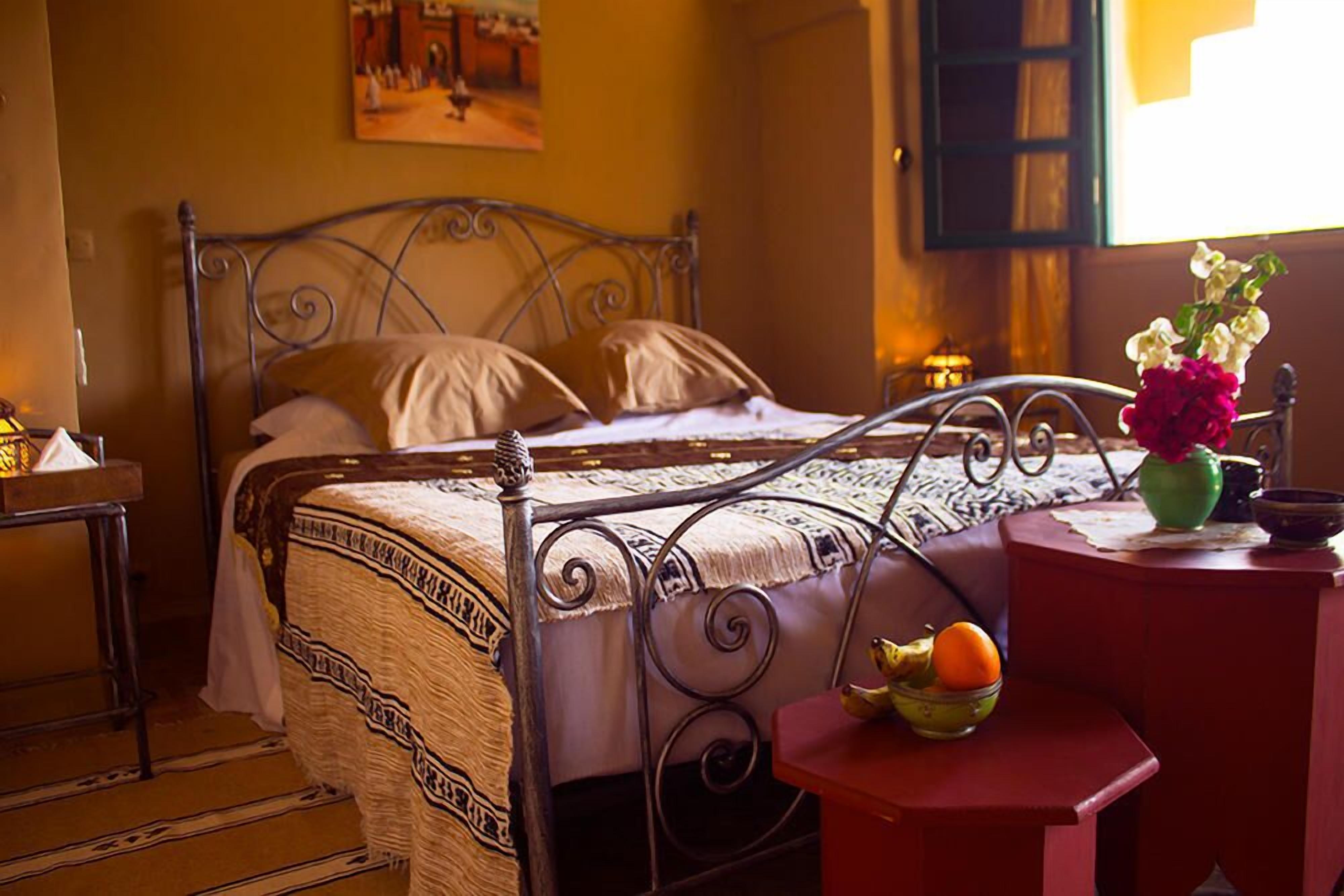 A bed with a metal frame, a bedside table with a vase of flowers, and a bowl of fruit.