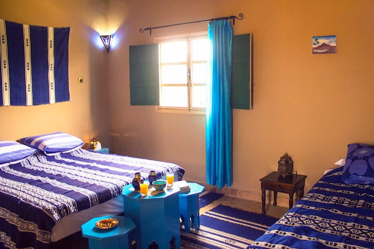 A room with two beds, a small table with a bowl of fruit and a glass of orange juice, a nightstand with a lamp, and a window with blue curtains.