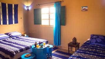 A room with two beds, a small table with a bowl of fruit and a glass of orange juice, a nightstand with a lamp, and a window with blue curtains.