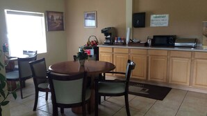 A dining area with a round table and chairs, a kitchen with a breakfast sign, and a window with natural light. - Executive Inn & Suites Schulenburg (Schulenburg)