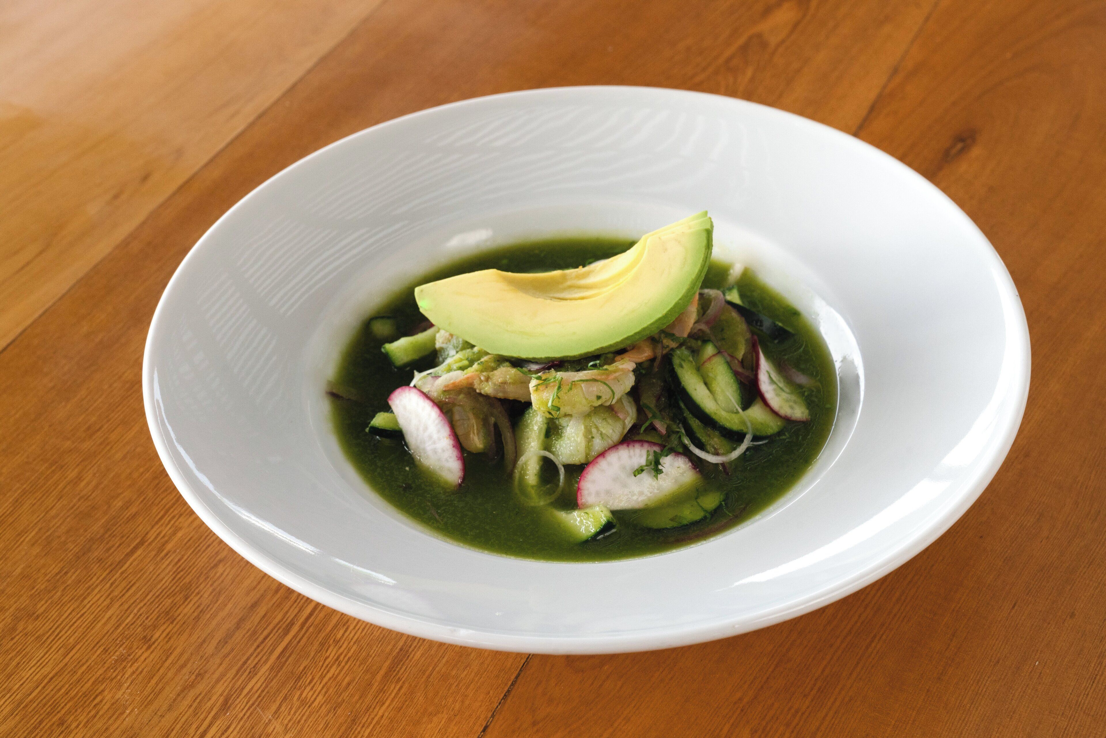 A white bowl with a green sauce, avocado, radishes, and cucumber slices on a wooden table.