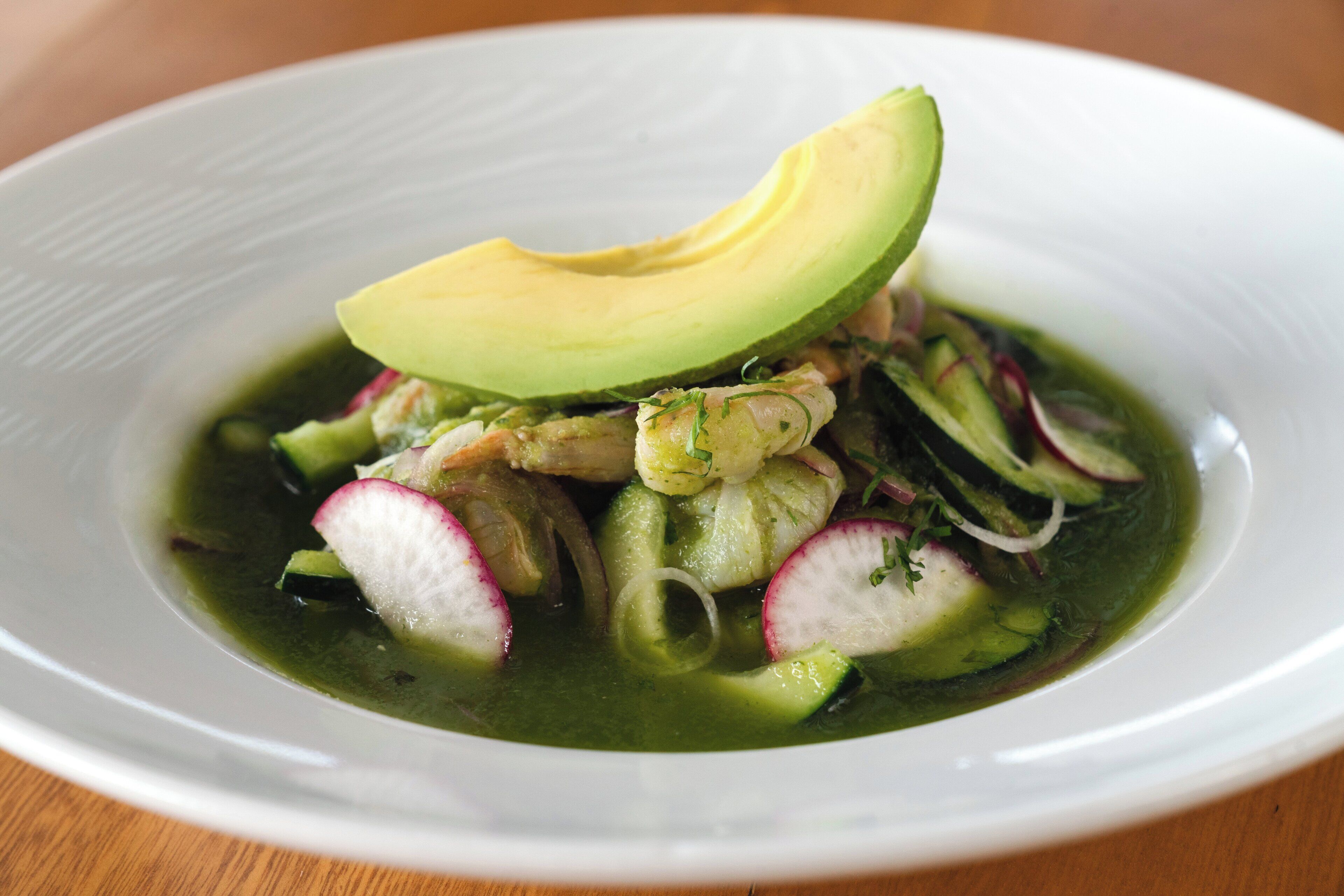 A white plate with a green vegetable soup, avocado, radishes, and cucumber slices.