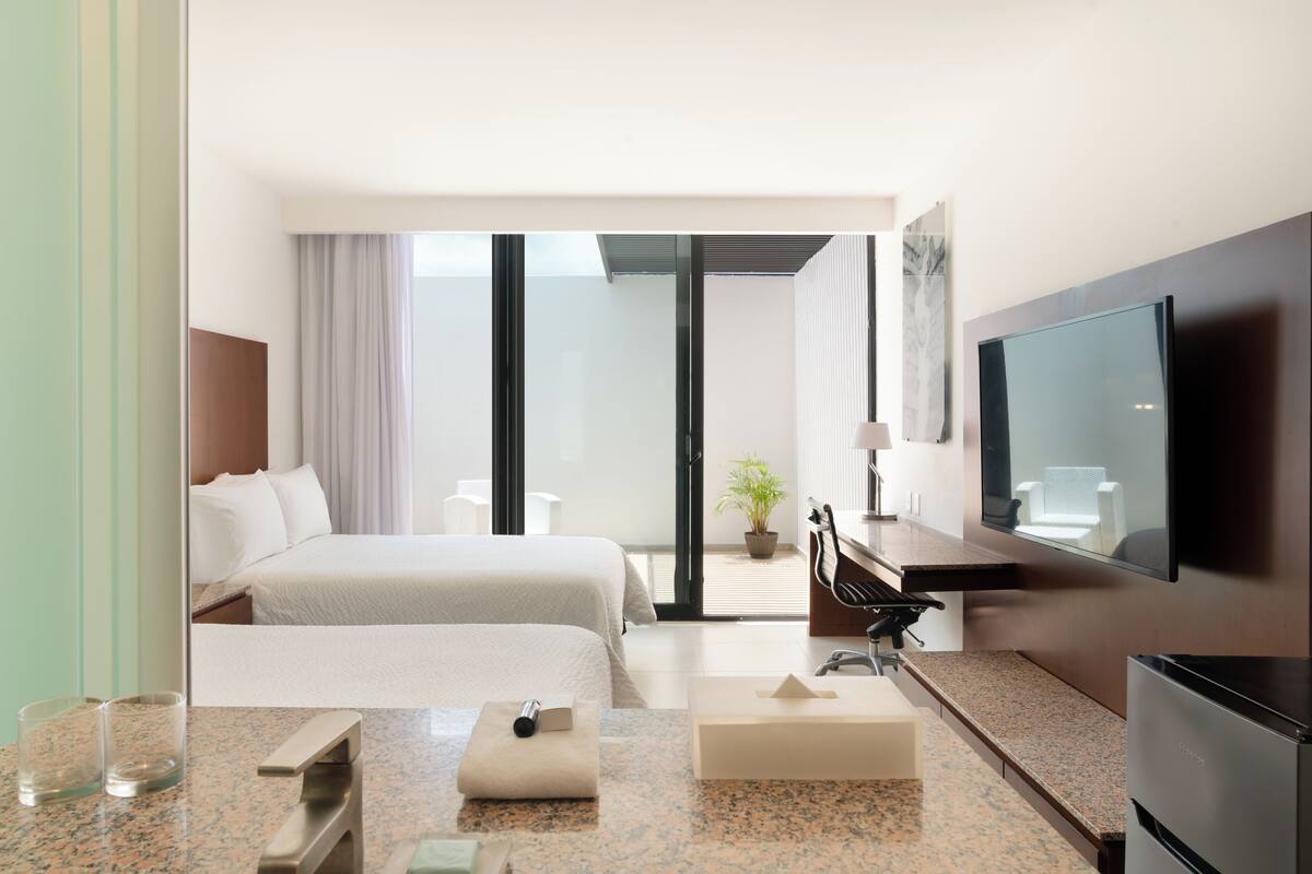Habitación de hotel con dos camas, un televisor de pantalla plana, un escritorio con una silla, una planta en maceta y un ventanal corredero que da a un balcón.