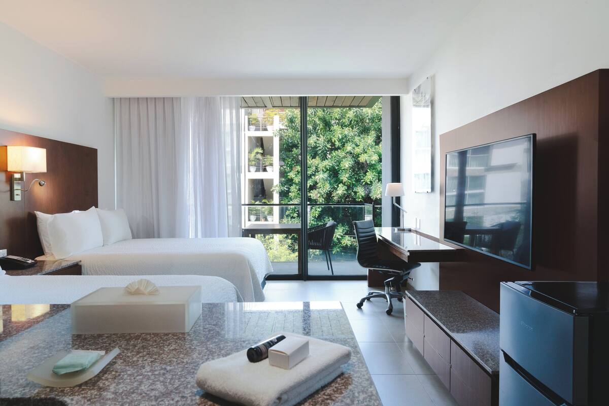 Habitación de hotel con cama, escritorio, televisor y balcón con vistas a la vegetación.