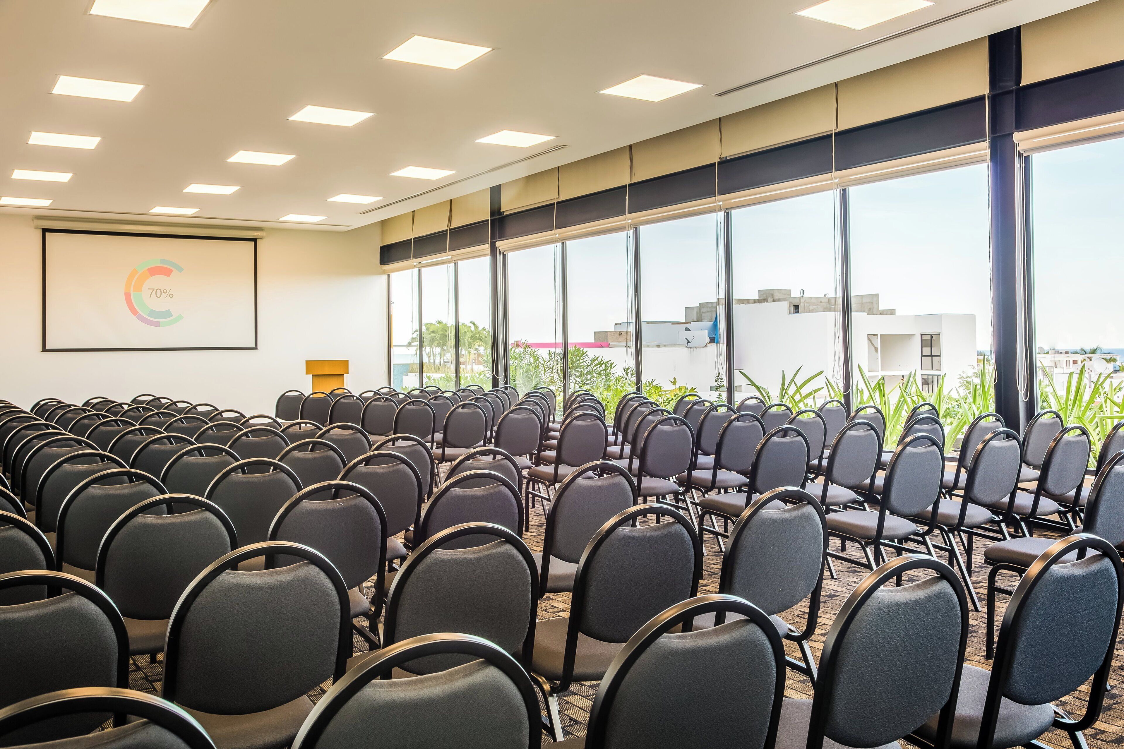 Un salón de conferencias con filas de sillas, amplias ventanas y una pantalla de proyector mostrando un porcentaje.