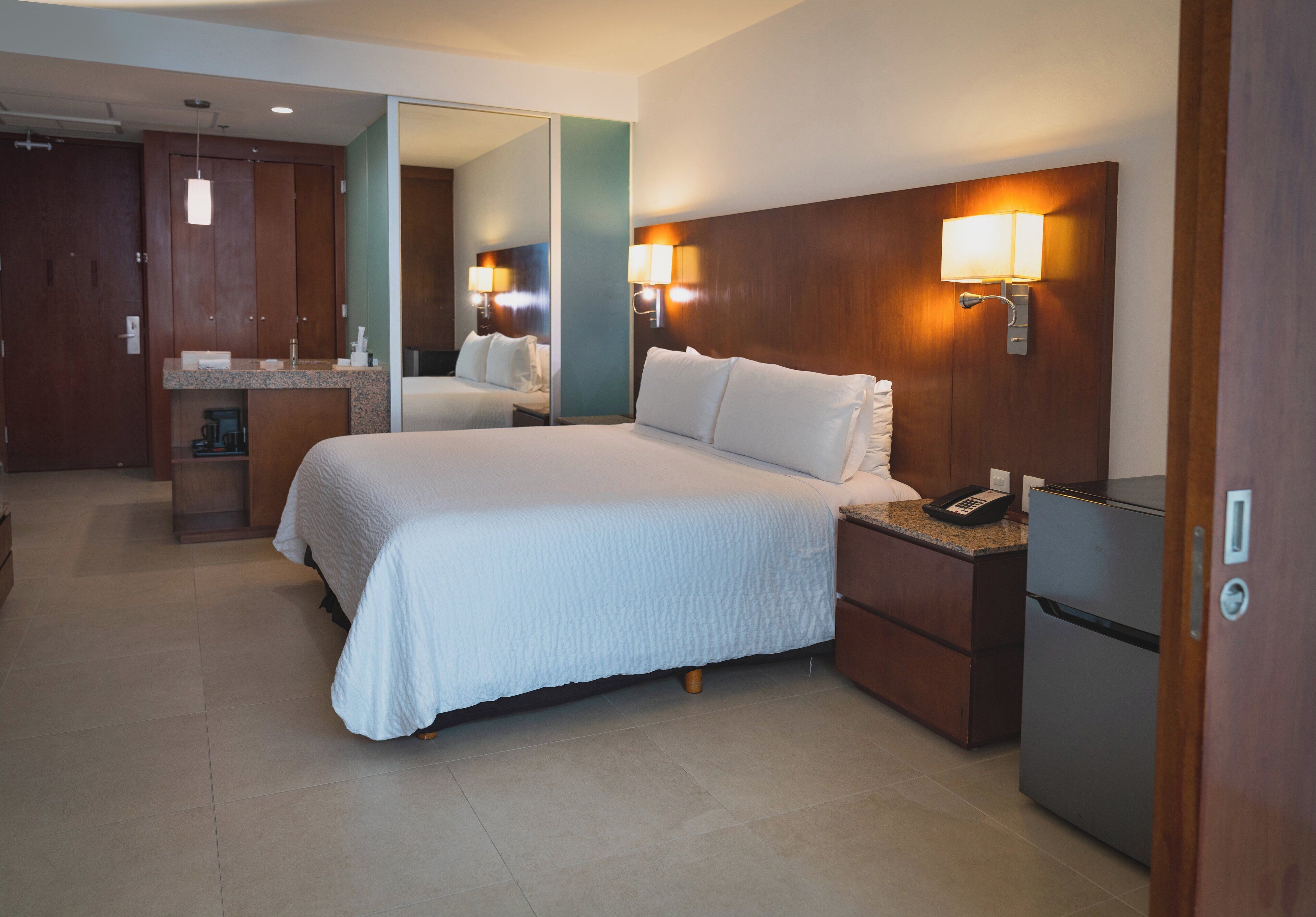 A hotel room with a large bed, bedside lamps, a small refrigerator, and a mirror.