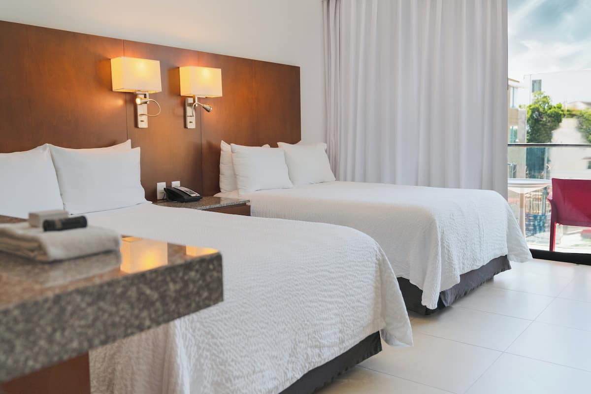 Two single beds with white bedspreads and pillows, situated against a headboard with wall-mounted lamps and a bedside table.