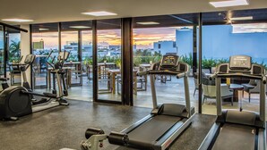 A gym with various exercise equipment, large windows, and a view of the cityscape at sunset. - Fiesta Inn Playa del Carmen (Playa del Carmen)