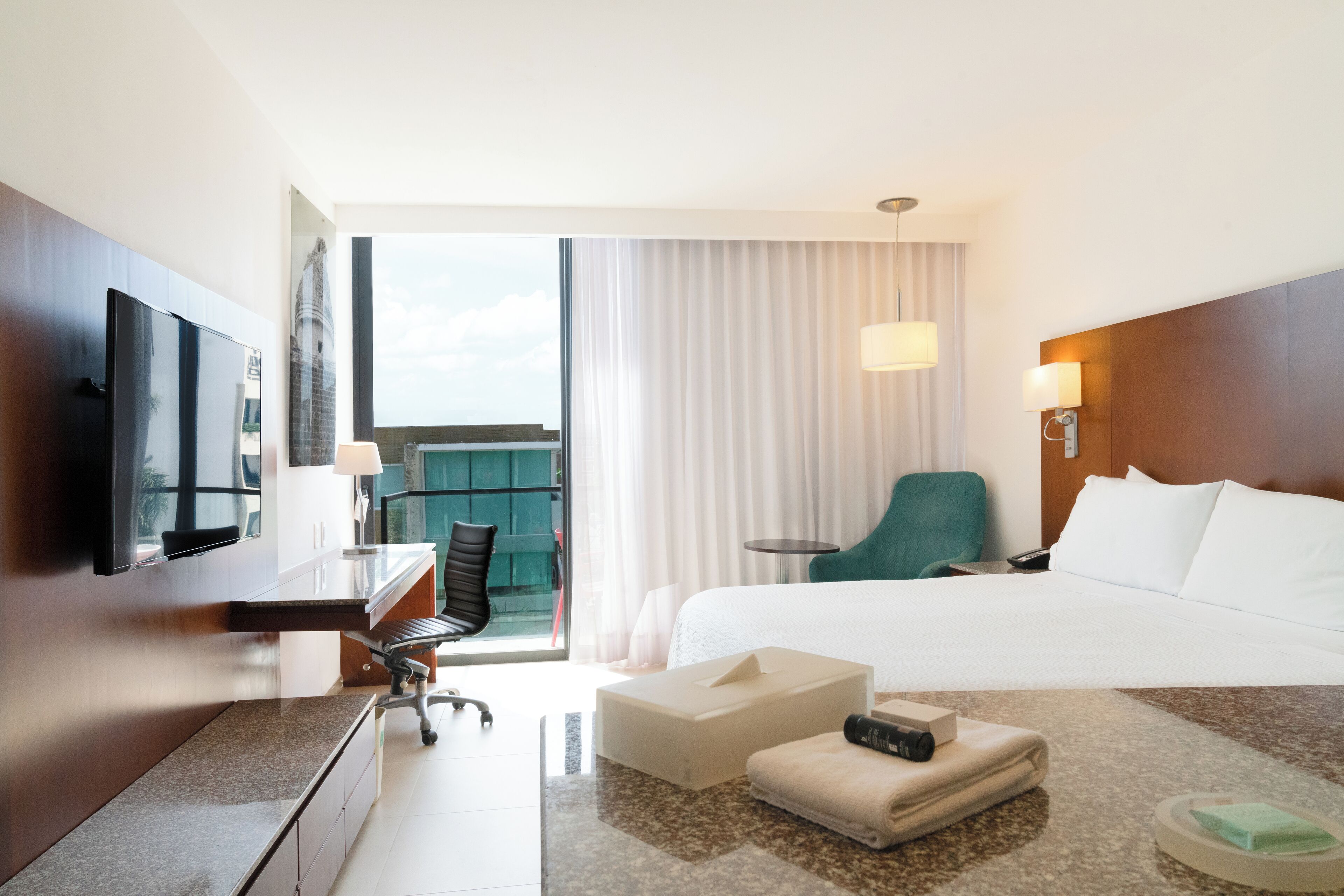 Ein Hotelzimmer mit einem Bett, einem Schreibtisch mit Stuhl, einem Fernseher, einem Fenster mit Vorhängen und einem Balkon mit Blick.