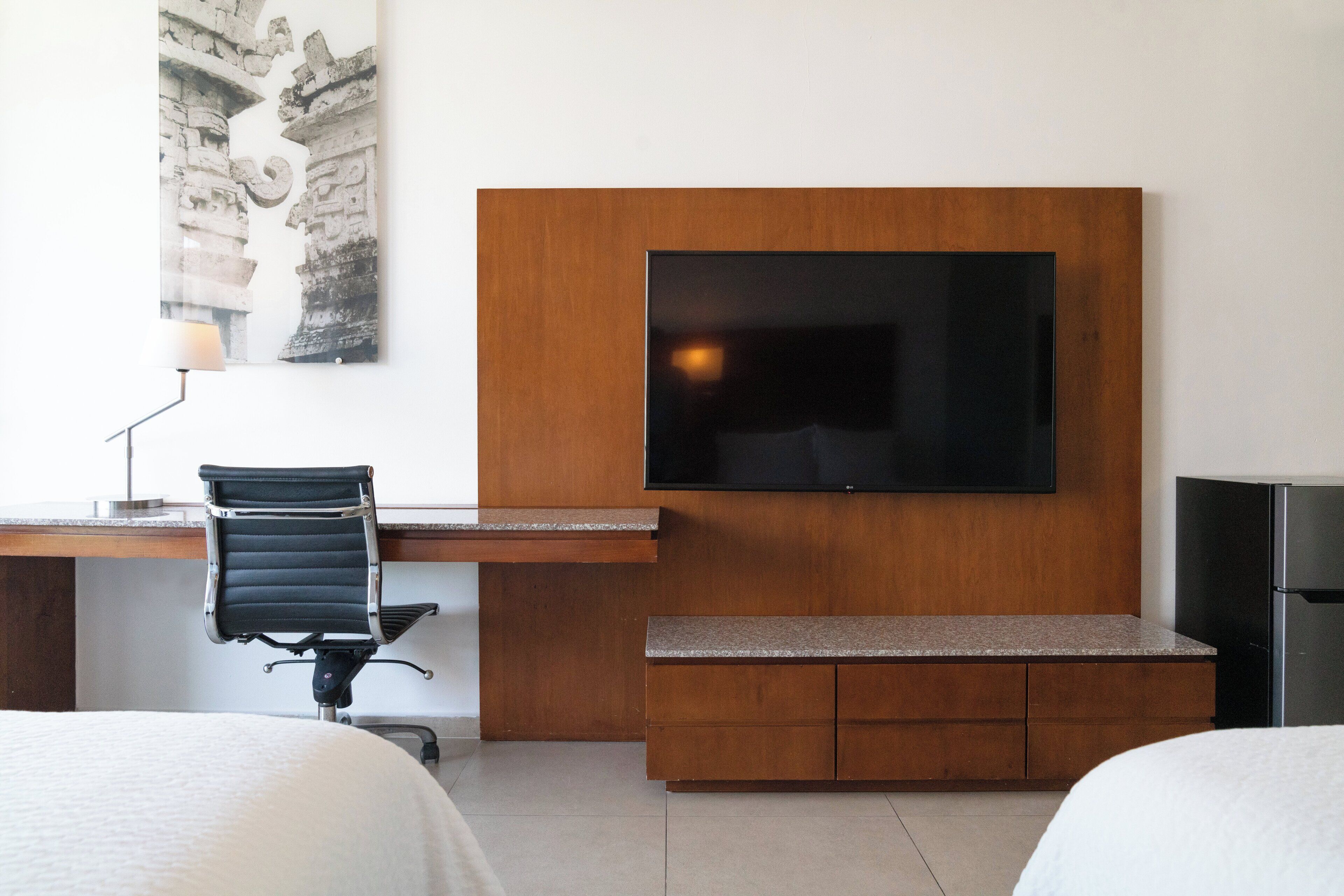 A hotel room with a wooden desk, a black office chair, a flat-screen TV mounted on the wall, a bench, and a bed.
