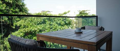 Una mesa de madera con una taza y platillo blancos, dos sillas negras y una barandilla de cristal con vistas a un paisaje verde exuberante.