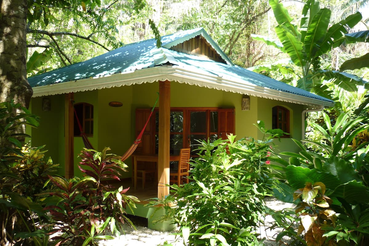 A small, single-story house with a green roof, surrounded by lush tropical vegetation.
