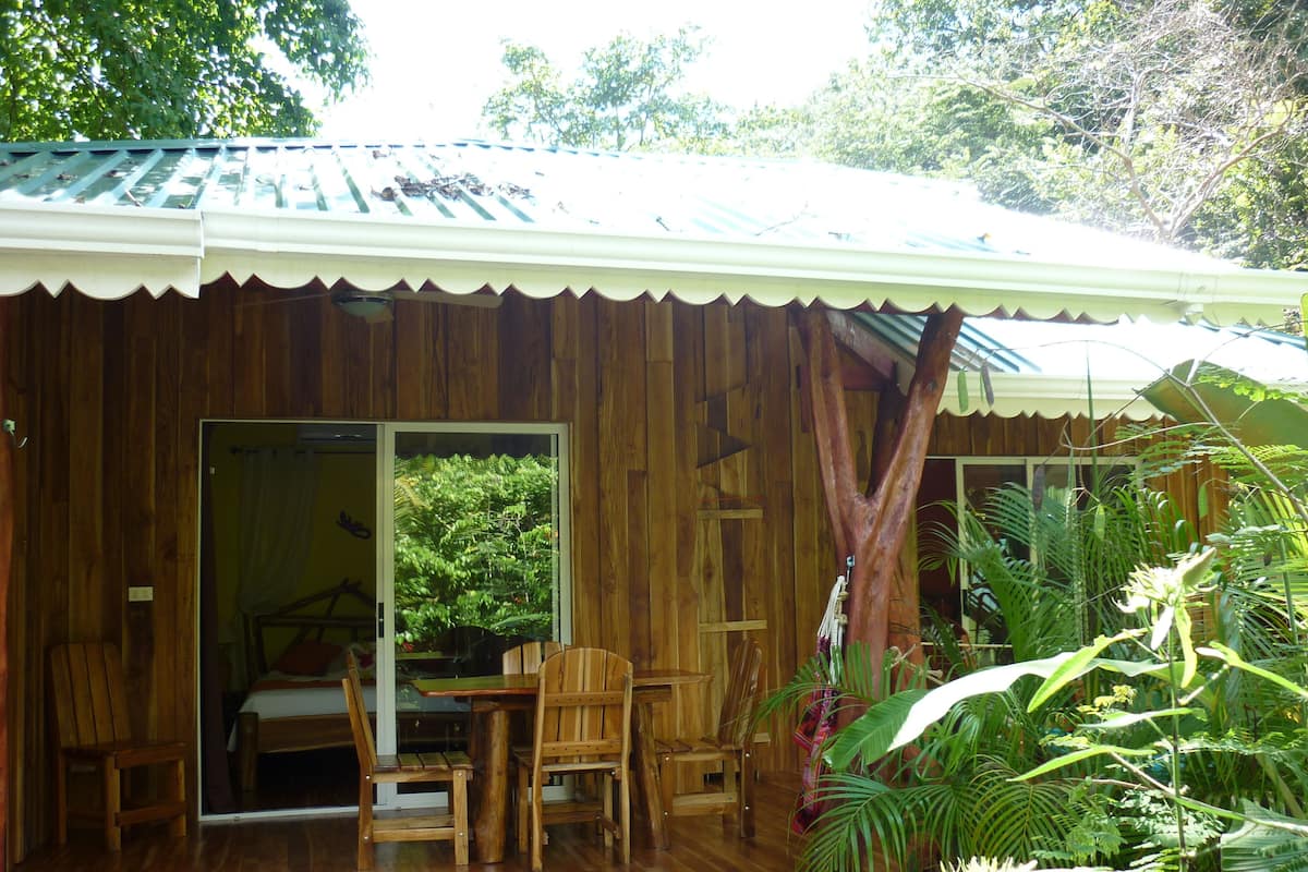 A wooden cabin with a covered porch, a dining area with chairs, and a view of lush greenery outside.