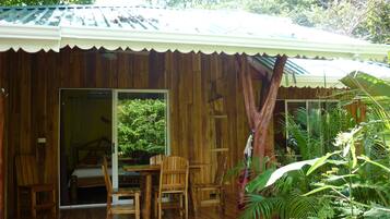 A wooden cabin with a covered porch, a dining area with chairs, and a view of lush greenery outside.