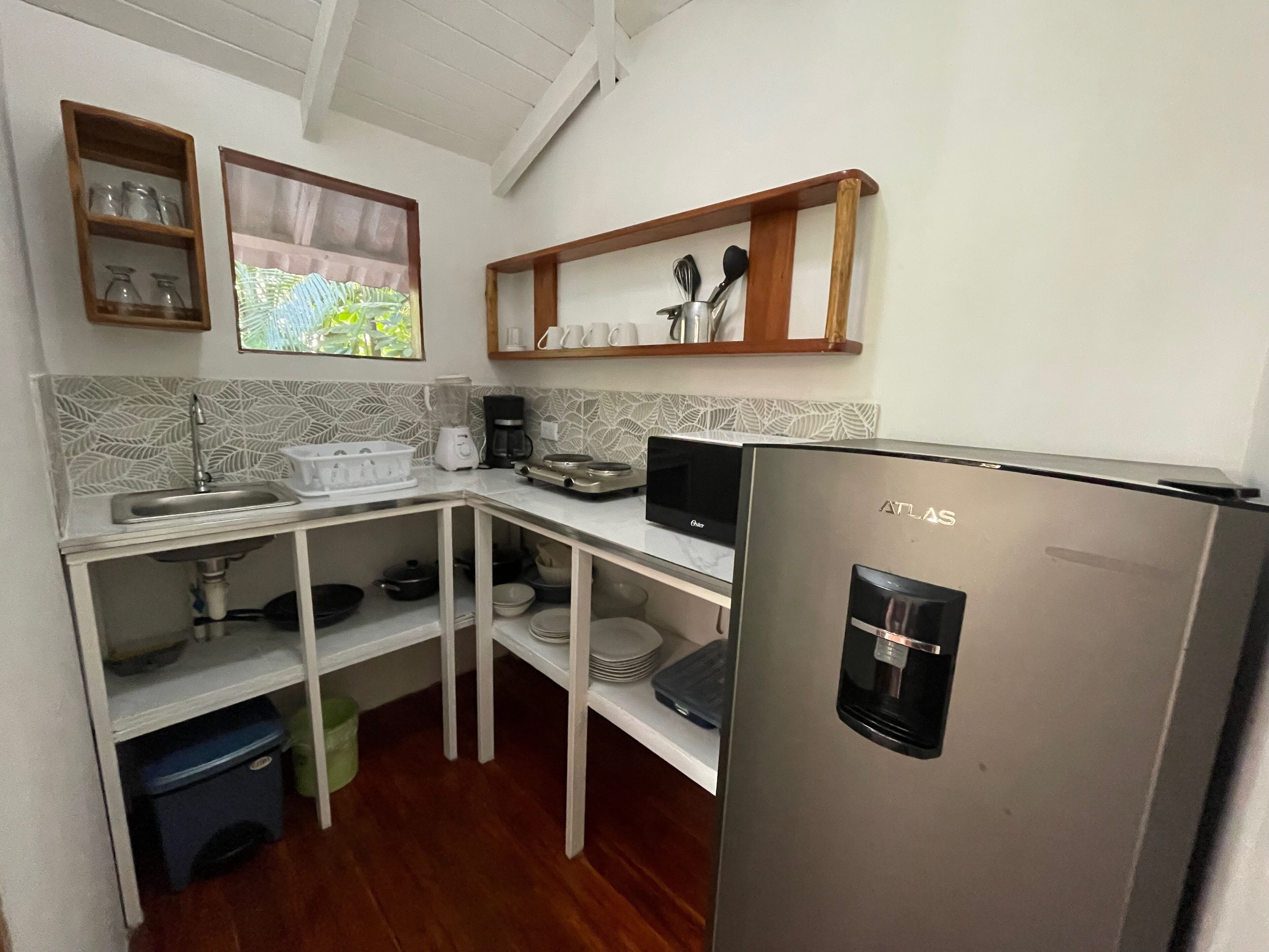 A compact kitchen with a built-in microwave, a coffee maker, and a refrigerator. There are shelves with dishes and a window with a view of greenery.
