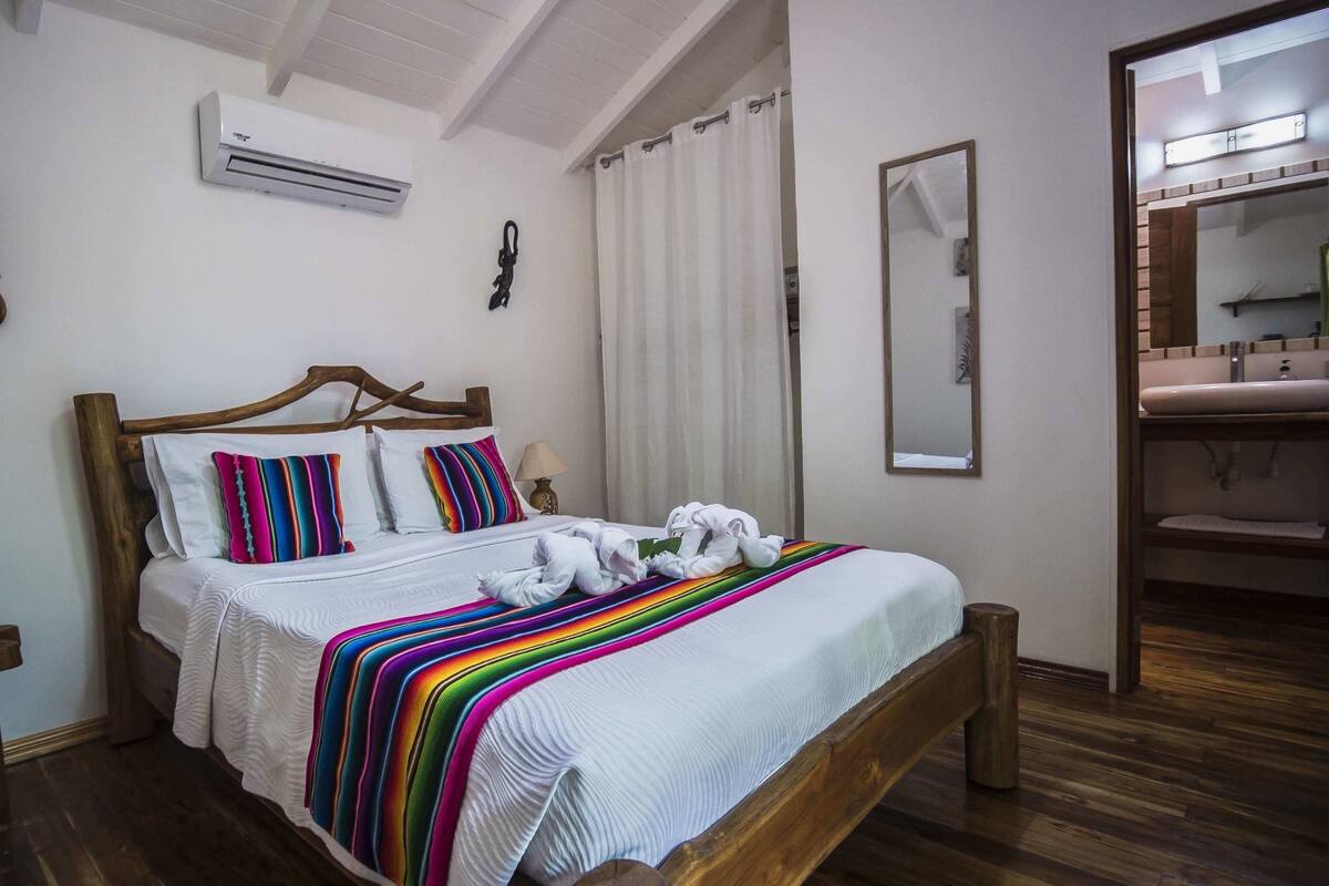 A neatly made bed with a colorful striped blanket, a wooden headboard, and a white wall with an air conditioning unit.