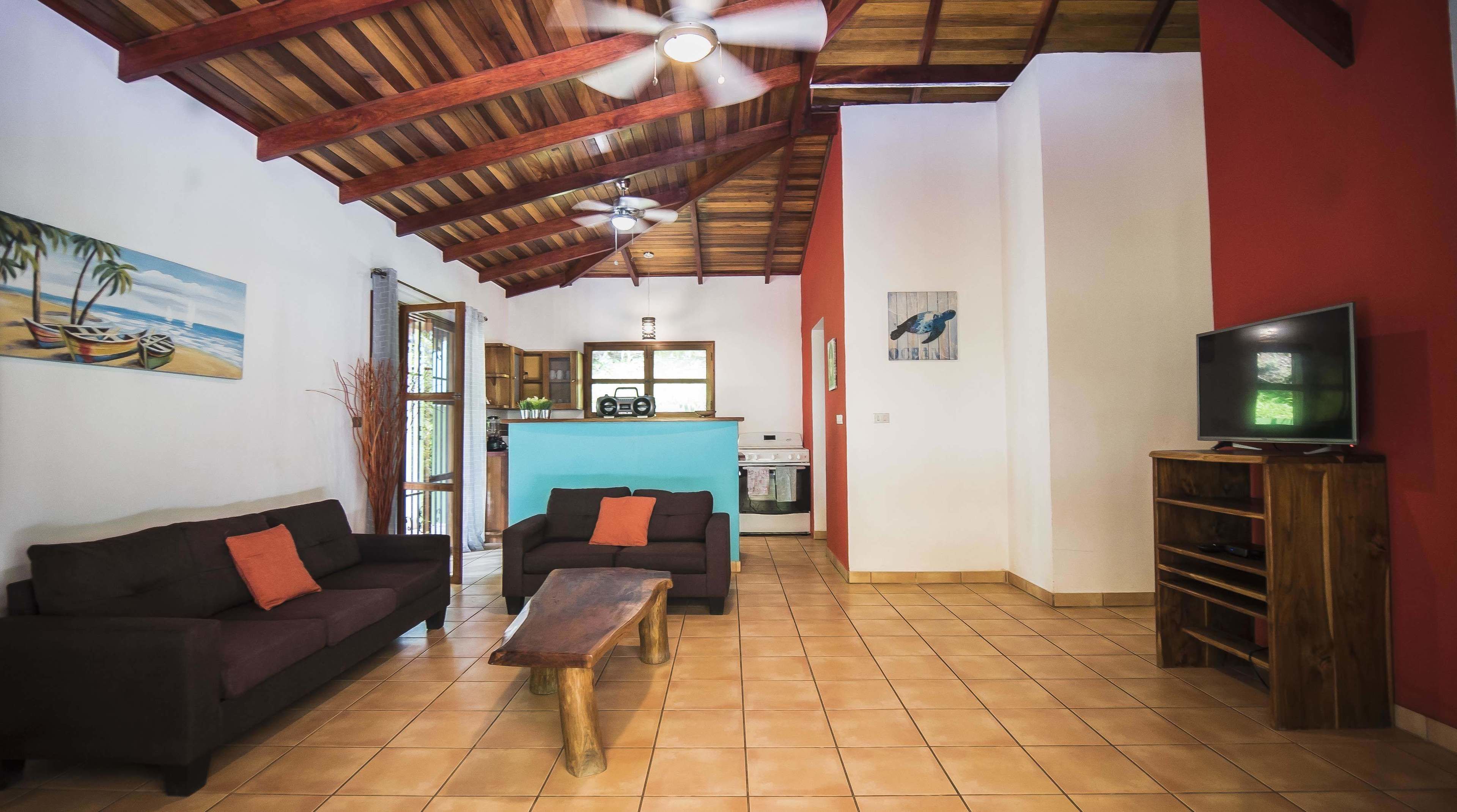 A living room with a sofa, a coffee table, a TV on a wooden stand, and a kitchen in the background.