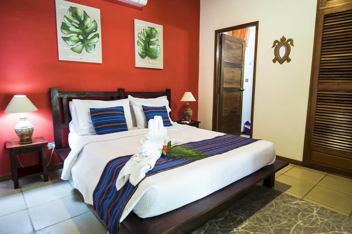 A hotel room with a bed, a nightstand with a lamp, a wooden door, and a wall with two leaf paintings.