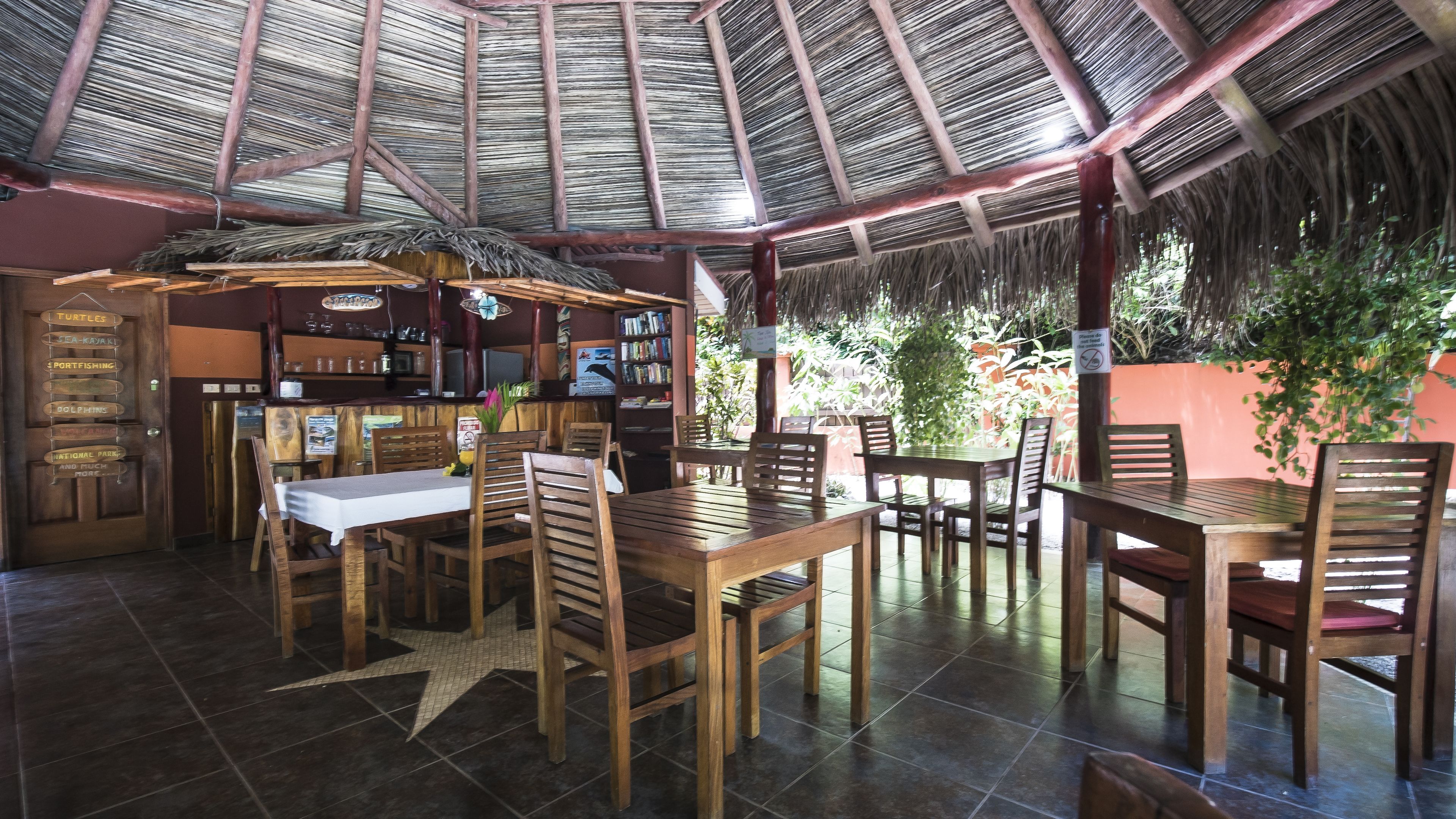 A restaurant with wooden tables and chairs, a thatched roof, and a bar area with shelves and a signboard.
