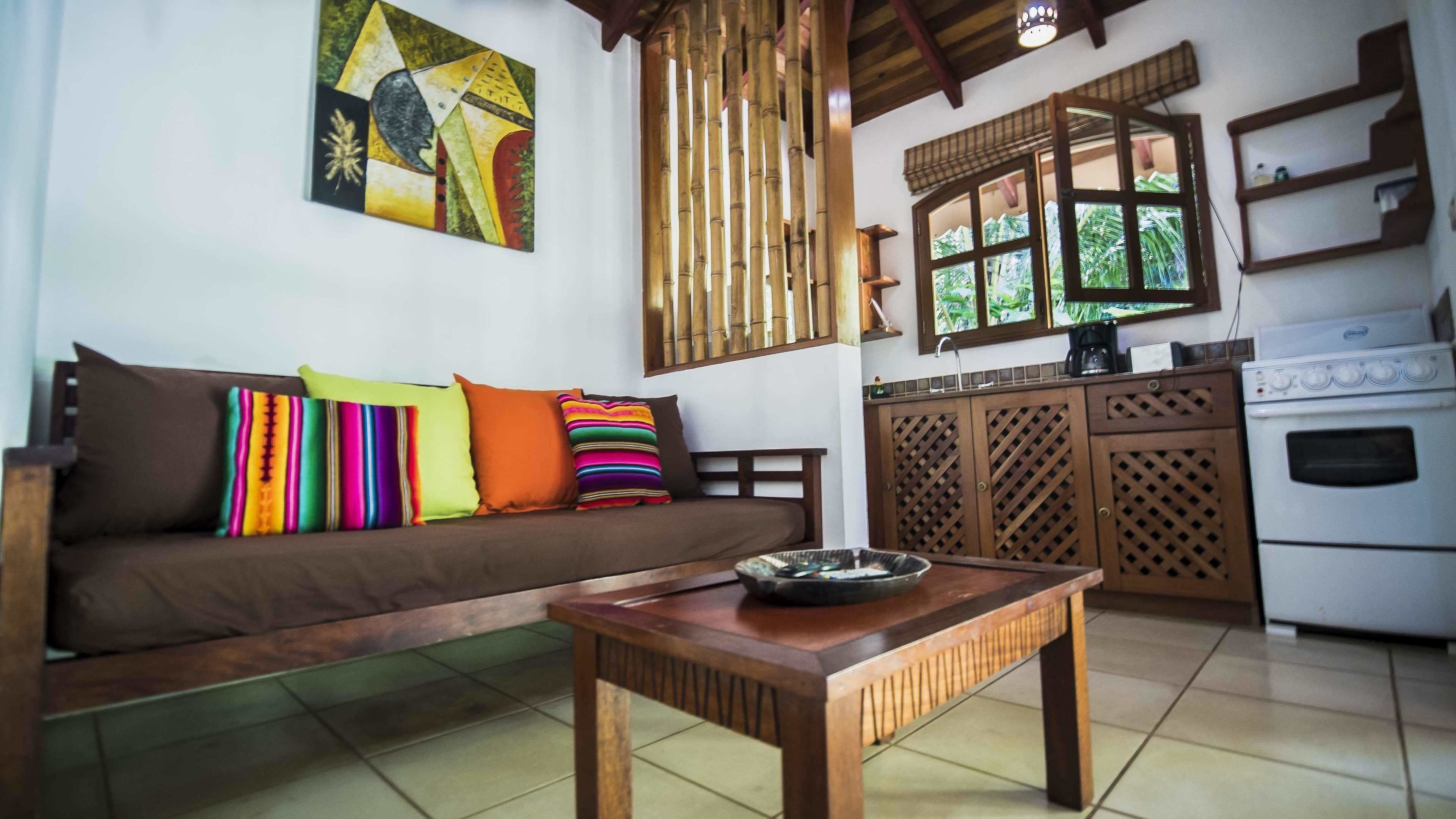 A living room with a brown sofa, colorful pillows, a wooden coffee table, a kitchen area with a stove, and a bamboo screen.