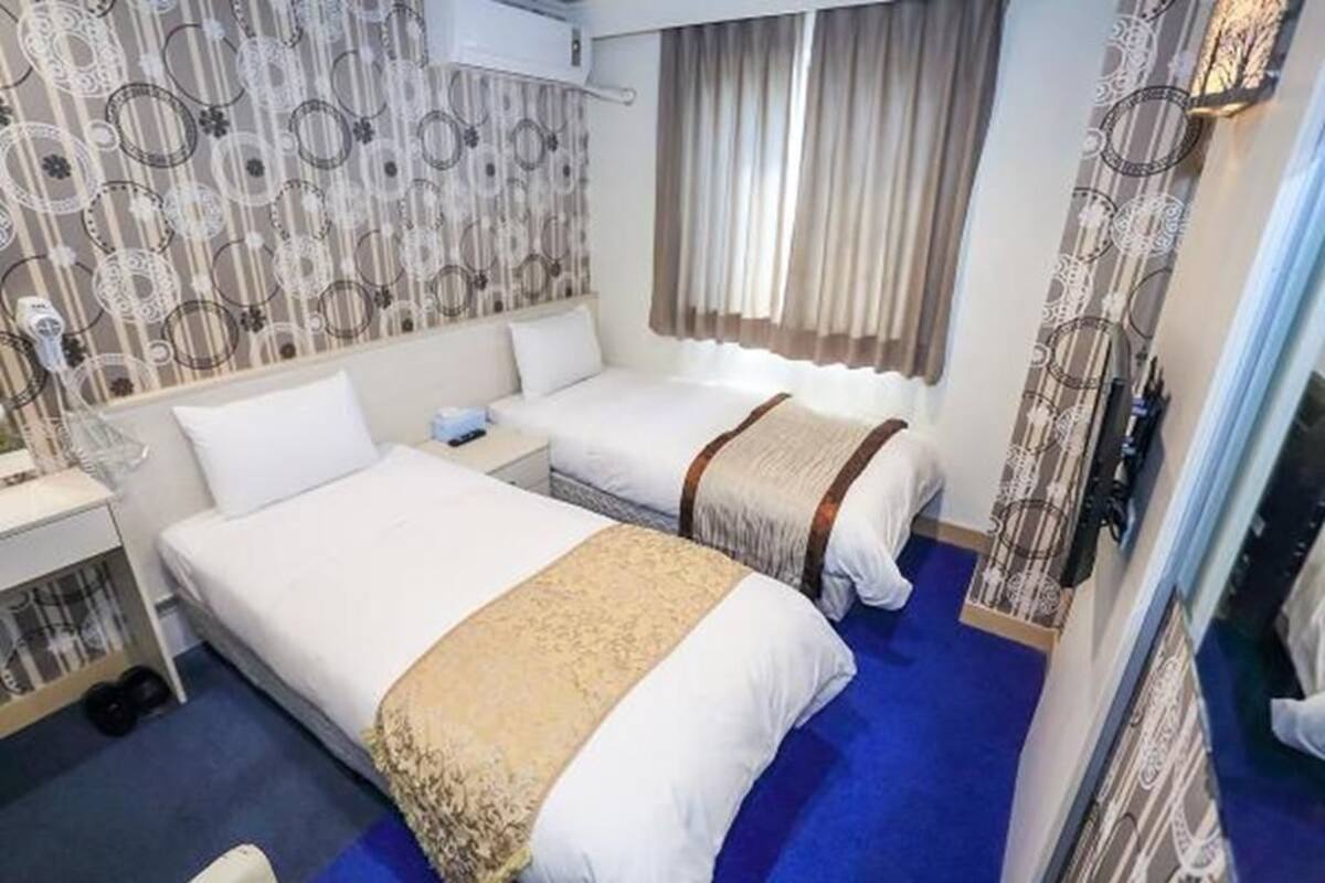 A hotel room with two beds, a window with curtains, a patterned wall, and a blue carpet.