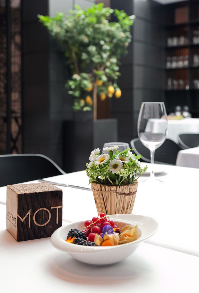 A dining table with a white plate of fruit salad, a wooden block with 'MOT' on it, and a woven basket with flowers.