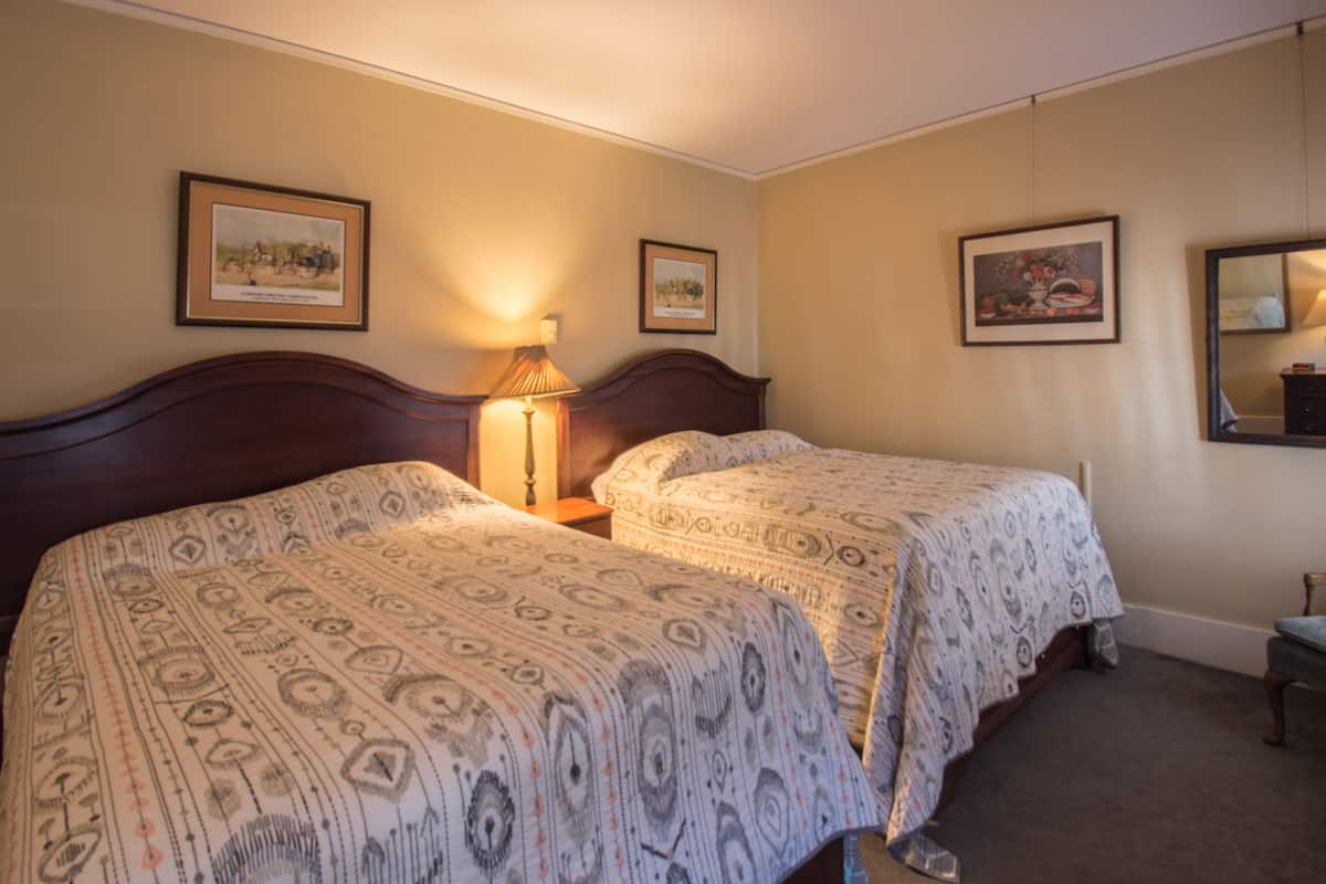 A hotel room with two single beds, a nightstand with a lamp, framed artwork on the walls, and a small table with a chair.