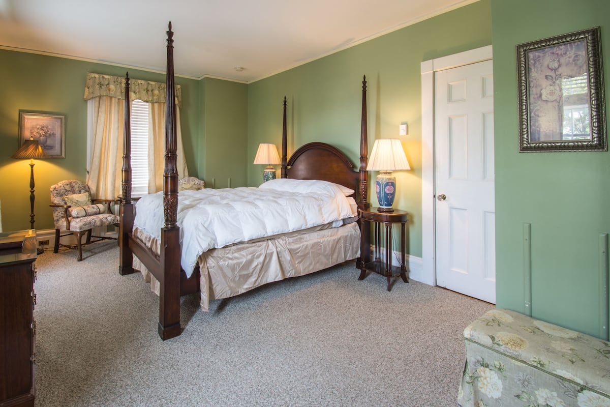 A four-poster bed with a wooden headboard, a nightstand with a lamp, a floral-patterned chair, and a door with a decorative frame.