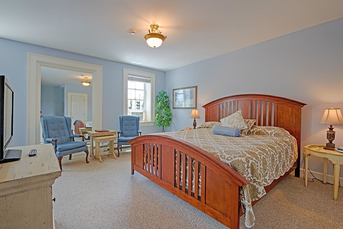 A bedroom with a wooden bed, two chairs, a desk, a lamp, and a television.