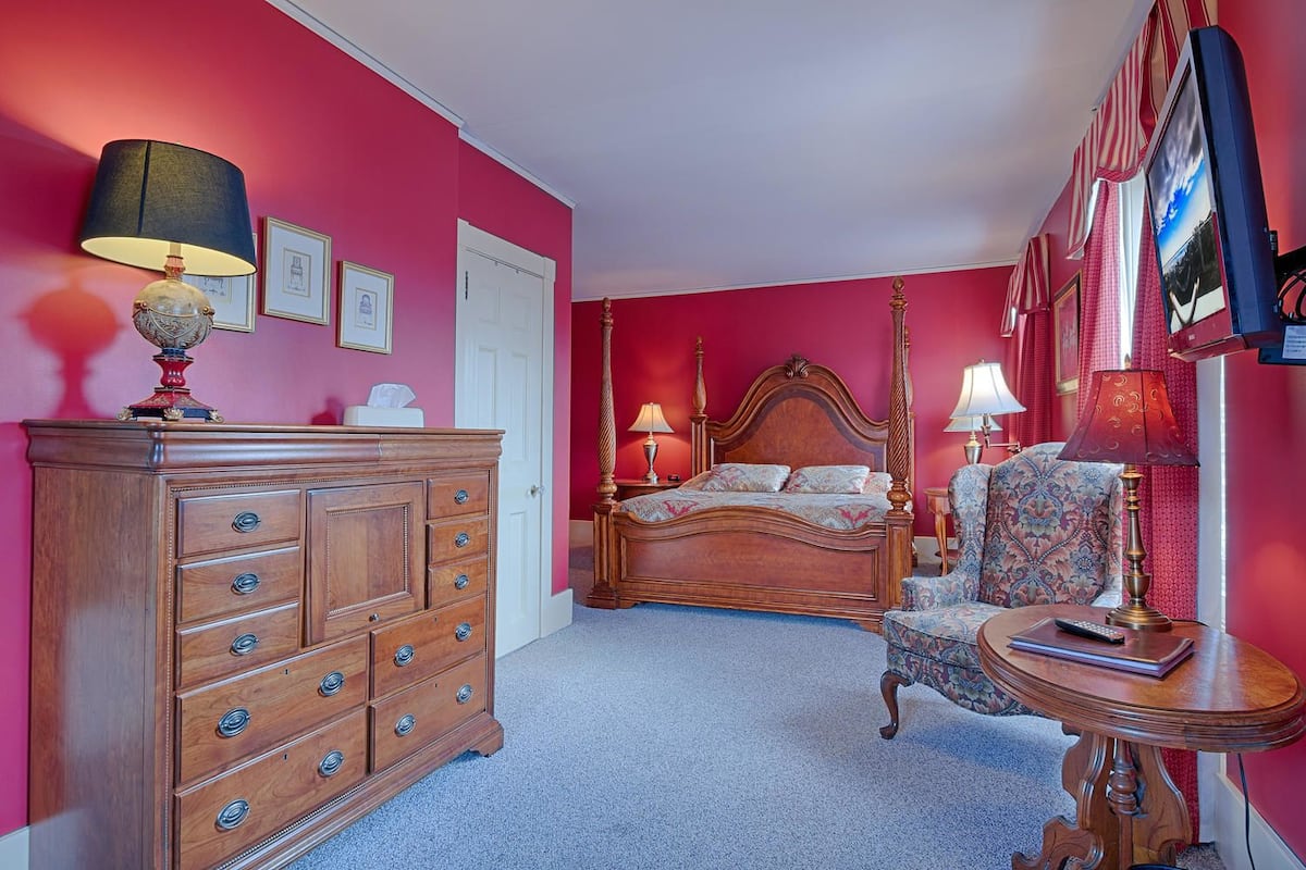 A bedroom with a four-poster bed, a wooden dresser, and a chair with a table.