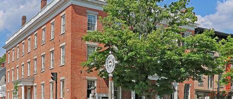 A historic brick building with a large tree in front, a sign indicating it is a historic site, and a bench along the sidewalk.